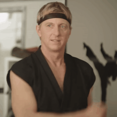 A Man Wearing A Black Karate Uniform And A Headband Is Standing In Front Of A Statue Of A Ninja . GIF