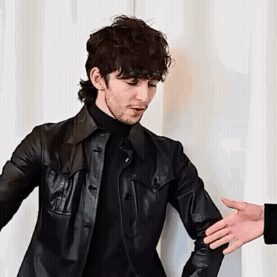 A Man Wearing A Black Leather Jacket And A Black Turtleneck Is Reaching Out To Shake Another Man 'S Hand . GIF