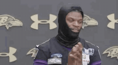 A Man Wearing A Black Mask And A Purple Jersey Is Standing In Front Of A Wall With Ravens Logos . GIF