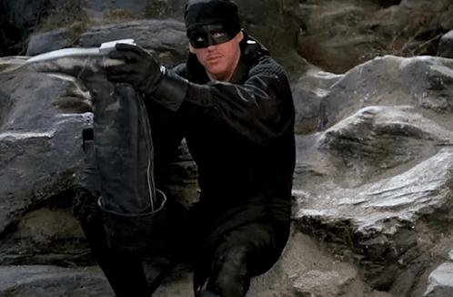 A Man Wearing A Black Mask And Gloves Is Sitting On A Rock GIF