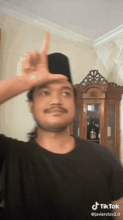A Man Wearing A Black Shirt And A Black Hat Is Making A Funny Face With His Finger On His Forehead .. GIF