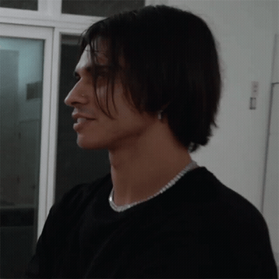A Man Wearing A Black Shirt And A Silver Necklace Looks To The Side GIF