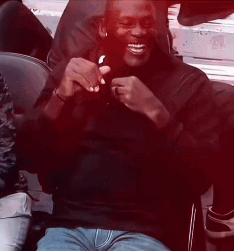 A Man Wearing A Black Shirt And Blue Jeans Is Sitting In A Chair And Smiling GIF