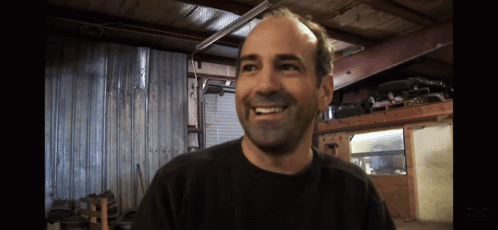 A Man Wearing A Black Shirt Is Smiling In A Garage GIF