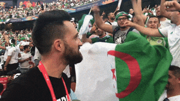 A Man Wearing A Black Shirt That Says ' A ' On It Stands In A Crowd Holding A Flag GIF