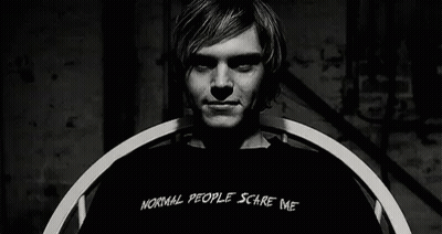 A Man Wearing A Black Shirt That Says `` Normal People Scare Me '' Is Sitting In A Chair . GIF