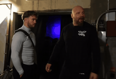 A Man Wearing A Black Shirt With A Butterfly On It Stands Next To Another Man Wearing A White Shirt GIF