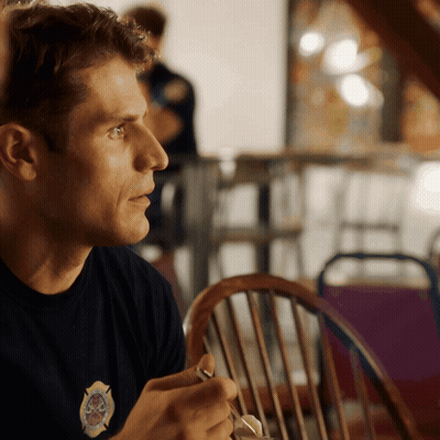 A Man Wearing A Black Shirt With A Firefighter Emblem On It Sits At A Table GIF
