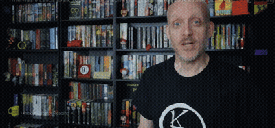 A Man Wearing A Black Shirt With A K On It Stands In Front Of A Bookshelf GIF