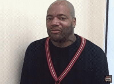 A Man Wearing A Black Shirt With A Red And White Striped Sweater Around His Neck Stands In Front Of A White Wall GIF