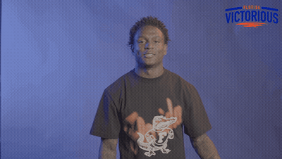 A Man Wearing A Black Shirt With An Alligator On It Stands In Front Of A Sign That Says Victorious GIF