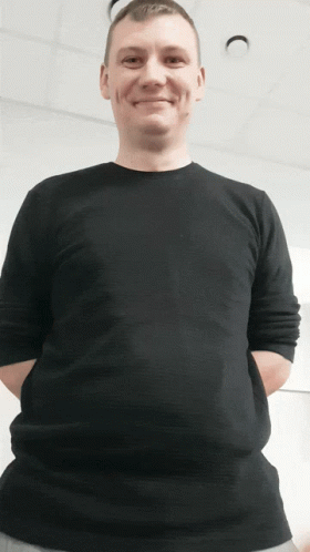 A Man Wearing A Black Shirt With His Hands In His Pockets Smiles GIF