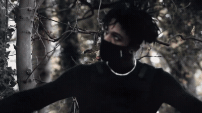A Man Wearing A Black Sweater And A Mask Is Standing In A Forest GIF