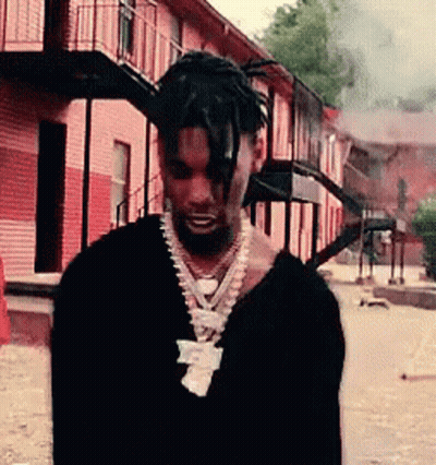 A Man Wearing A Black Sweater And A Necklace Is Standing In Front Of A Building . GIF