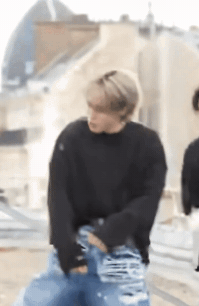 A Man Wearing A Black Sweater And Blue Jeans Is Standing On A Sidewalk . GIF