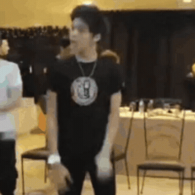 A Man Wearing A Black T-Shirt And A Necklace Is Standing In A Room . GIF