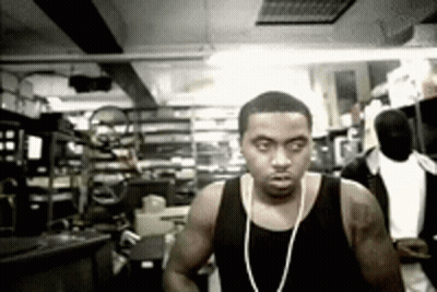 A Man Wearing A Black Tank Top And A Necklace Stands In A Room GIF