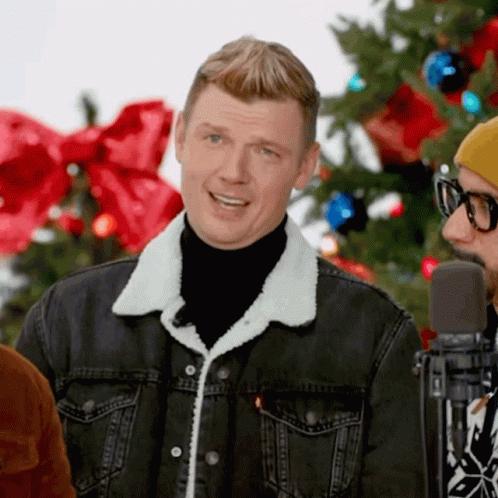 A Man Wearing A Black Turtleneck And A Denim Jacket Stands In Front Of A Microphone In Front Of A Christmas Tree GIF