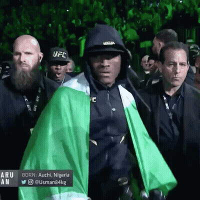 A Man Wearing A Black Ufc Hat And A Green Cape Walks Through A Crowd GIF