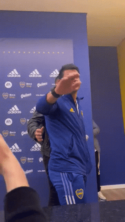 A Man Wearing A Blue Adidas Jacket Stands In Front Of A Blue Wall GIF