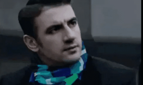 A Man Wearing A Blue And Green Scarf Around His Neck Looks At The Camera . GIF