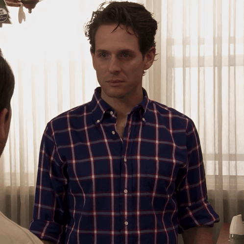 A Man Wearing A Blue And Red Plaid Shirt Is Standing In Front Of A Window GIF