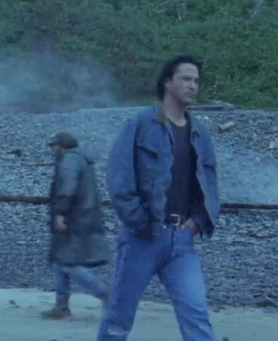 A Man Wearing A Blue Denim Jacket And Jeans Is Walking On A Rocky Beach . GIF