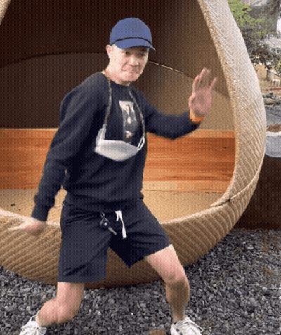 A Man Wearing A Blue Hat And A Face Mask Is Standing In Front Of A Wicker Chair GIF