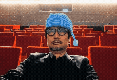 A Man Wearing A Blue Hat And Glasses Sits In A Theater GIF