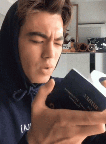 A Man Wearing A Blue Hoodie Is Reading A Book With The Word Bible On It GIF