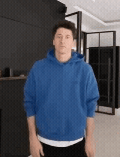 A Man Wearing A Blue Hoodie Is Standing In A Living Room . GIF