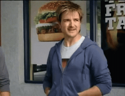A Man Wearing A Blue Hoodie Stands In Front Of A Mcdonald 'S Advertisement GIF