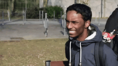 A Man Wearing A Blue Jacket And A Backpack Is Smiling While Standing In A Park . GIF