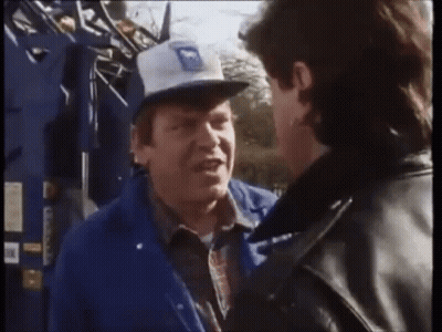 A Man Wearing A Blue Jacket And A White Hat With The Letter M On It Talks To Another Man GIF