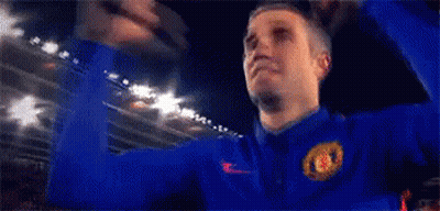 A Man Wearing A Blue Jacket With The Manchester United Logo On It Is Raising His Fist In The Air . GIF