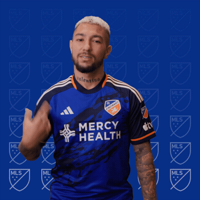 A Man Wearing A Blue Mercy Health Jersey Stands In Front Of A Blue Background With Mls Logos GIF
