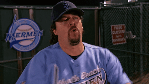 A Man Wearing A Blue Mermen Baseball Jersey Stands In Front Of A Fence GIF