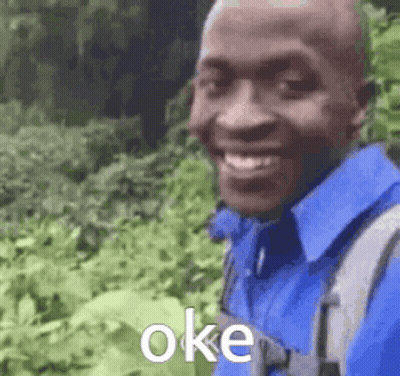 A Man Wearing A Blue Shirt And A Backpack Is Smiling And Standing In A Field . GIF