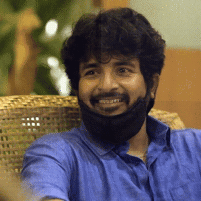 A Man Wearing A Blue Shirt And A Black Face Mask Smiles While Sitting In A Chair GIF