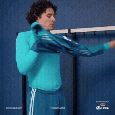 A Man Wearing A Blue Shirt And Blue Shorts Is Standing In A Locker Room With Corona Written On The Bottom GIF