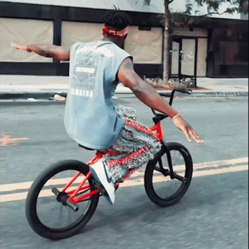A Man Wearing A Blue Shirt That Says Jamaica On The Back Is Riding A Bike GIF