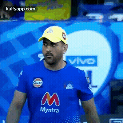 A Man Wearing A Blue T-Shirt And A Yellow Hat Is Standing In Front Of A Vivo Logo . GIF
