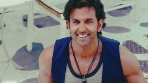 A Man Wearing A Blue Vest And A Necklace Smiles GIF