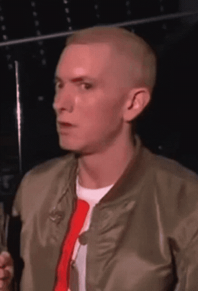 A Man Wearing A Bomber Jacket And A Red Tie Is Talking Into A Microphone GIF