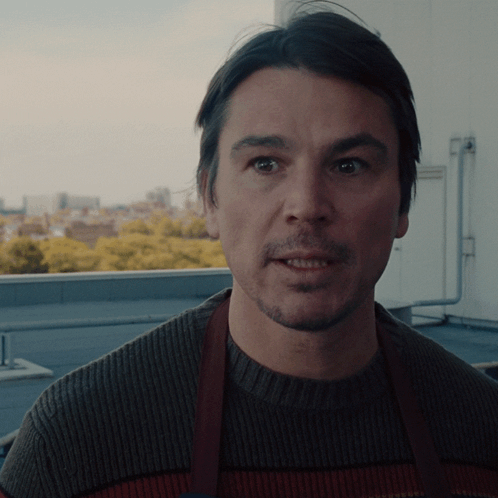 A Man Wearing A Brown Apron And A Sweater Looks At The Camera GIF