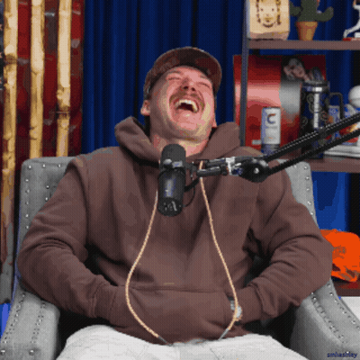 A Man Wearing A Brown Hoodie Is Laughing In Front Of A Microphone With The Letter C On It GIF