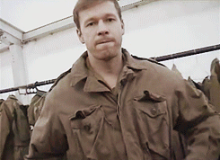 A Man Wearing A Brown Jacket Is Standing In Front Of A Rack Of Clothes GIF