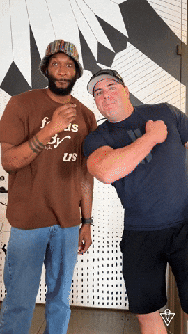 A Man Wearing A Brown Shirt That Says ' Foods By Us ' On It Stands Next To Another Man GIF