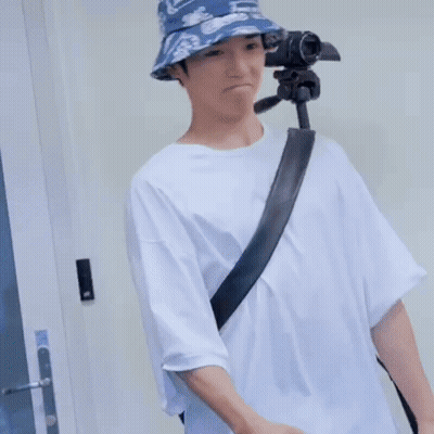 A Man Wearing A Bucket Hat And A White Shirt Is Standing Next To A Tripod . GIF