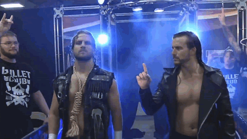 A Man Wearing A Bullet Club Shirt Stands Next To Another Man GIF